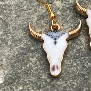 K&ouml;nnte beinhalten: Ein Paar goldfarbene Ohrringe mit wei&szlig;en und schwarzen Totenkopf-Anh&auml;ngern. Die Totenk&ouml;pfe haben ein geometrisches Muster und goldene Akzente.