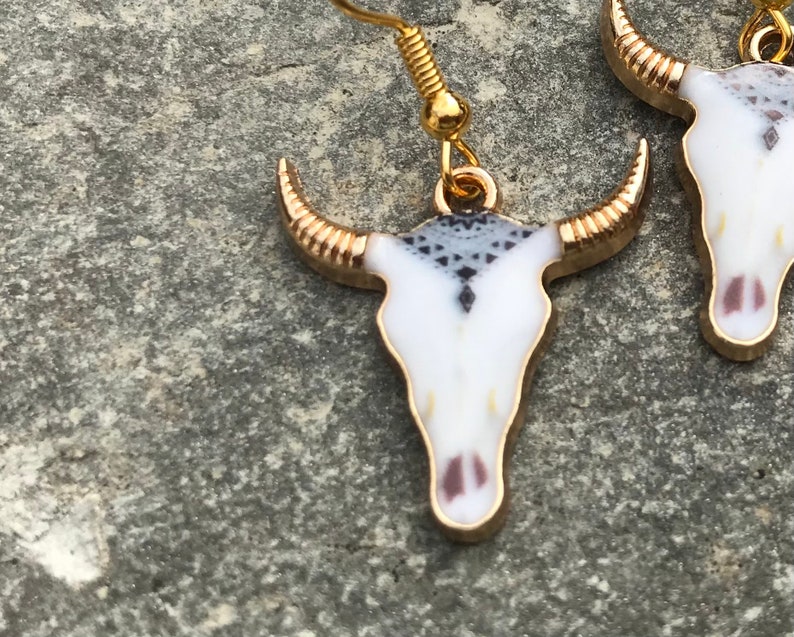 K&ouml;nnte beinhalten: Ein Paar goldfarbene Ohrringe mit wei&szlig;en und schwarzen Totenkopf-Anh&auml;ngern. Die Totenk&ouml;pfe haben ein geometrisches Muster und goldene Akzente.