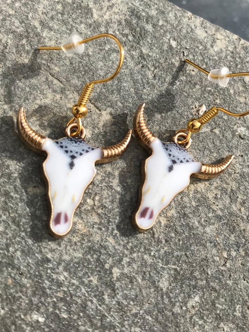 K&ouml;nnte beinhalten: Ein Paar goldfarbene Ohrringe mit einem wei&szlig;en und schwarzen Totenkopfdesign. Der Totenkopf hat ein detailliertes Muster und eine goldene Umrandung.