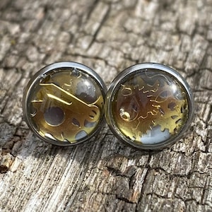 Puede incluir: Un par de pendientes de botón plateados con resina transparente y engranajes dorados en el interior.