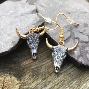 May include: A pair of gold tone earrings with a black and white patterned bull skull design.