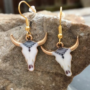 K&ouml;nnte beinhalten: Ein Paar goldfarbene Ohrringe mit einem wei&szlig;en und schwarzen Stierkopfdesign. Die Ohrringe sind im H&auml;nge-Stil mit einem Hakenverschluss.