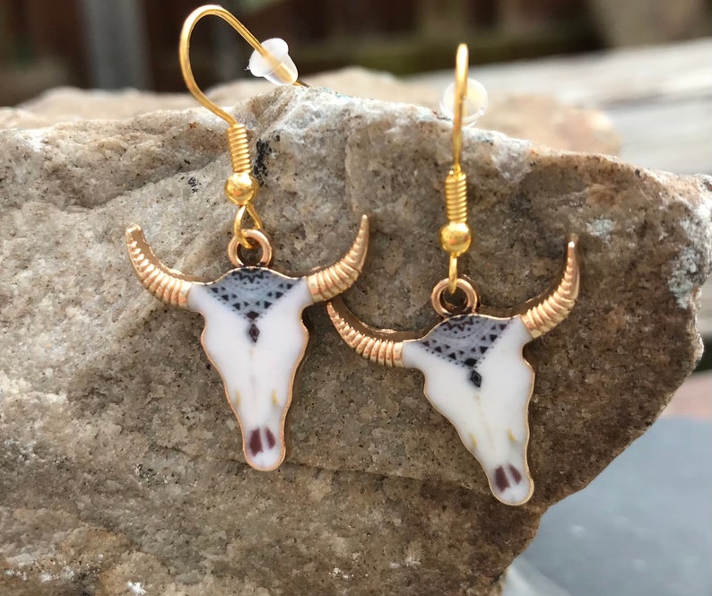 K&ouml;nnte beinhalten: Ein Paar goldfarbene Ohrringe mit einem wei&szlig;en und schwarzen Stierkopfdesign. Die Ohrringe sind im H&auml;nge-Stil mit einem Hakenverschluss.