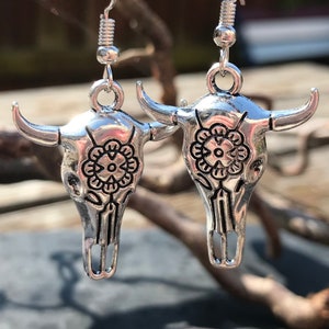May include: A pair of silver earrings featuring a detailed skull design with a floral pattern. The earrings are made of metal and have a dangling design.