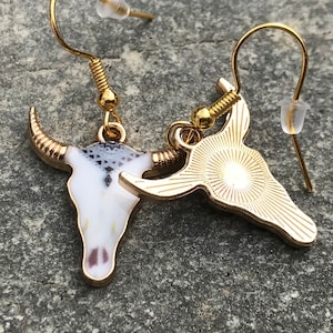 K&ouml;nnte beinhalten: Ohrringe mit goldfarbenem Anh&auml;nger, die ein Stierkopfdesign zeigen. Ein Ohrring hat einen wei&szlig;en und schwarzen Emaille-Sch&auml;del, w&auml;hrend der andere aus massivem Goldton ist.