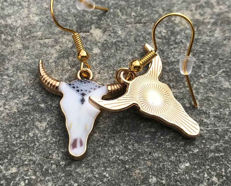 K&ouml;nnte beinhalten: Ohrringe mit goldfarbenem Anh&auml;nger, die ein Stierkopfdesign zeigen. Ein Ohrring hat einen wei&szlig;en und schwarzen Emaille-Sch&auml;del, w&auml;hrend der andere aus massivem Goldton ist.