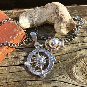 Op de afbeelding: Een zilveren ketting met een kompas hanger en een kleine globe bedel. De ketting is gemaakt van een zilveren ketting en de kompas hanger heeft een stervormig ontwerp in het midden.