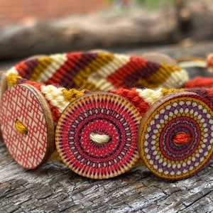 May include: A woven friendship bracelet with three round wooden beads. The beads are decorated with geometric patterns in red, orange, yellow, and purple.