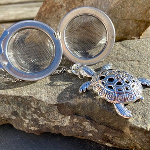 May include: A silver tea infuser with a turtle-shaped charm. The infuser is open, revealing a fine mesh basket. The turtle charm is detailed with a patterned shell and outstretched flippers, connected to the infuser by a chain.