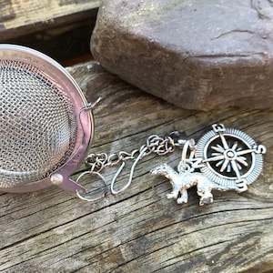 May include: Silver tea infuser with a chain, compass charm, and a small animal charm. The infuser is on a wooden surface.