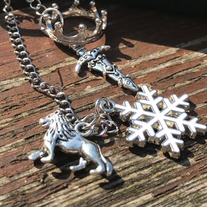 May include: A silver chain necklace with four charms: a crown, a sword, a snowflake, and a lion. The charms are all silver and have a detailed design.