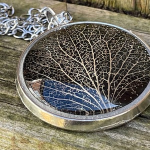 May include: A silver pendant necklace with a round resin charm featuring a delicate, gold-toned leaf skeleton. The leaf is encased in clear resin and has a black background.