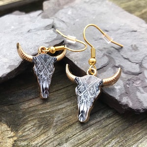 May include: A pair of gold dangle earrings with a black and white bull skull design. The skull has a geometric pattern.