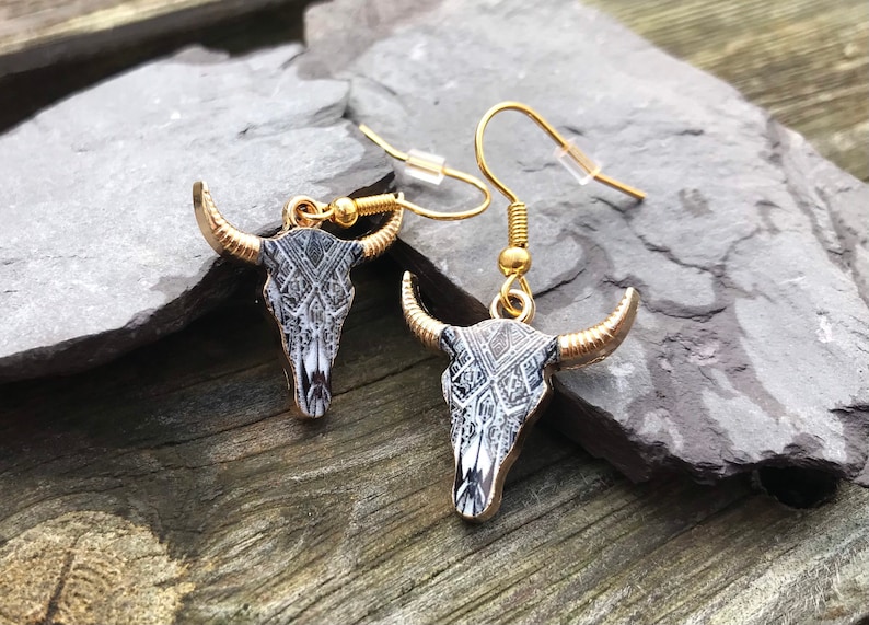 May include: A pair of gold dangle earrings with a black and white bull skull design. The skull has a geometric pattern.