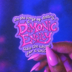 May include: A die-cut sticker with a purple background and pink lettering that reads: On the verge of letting a DEMONIC ENTITY take the wheel for a while. The text features a stylised, bold font with a glossy, reflective appearance and dark outlines.