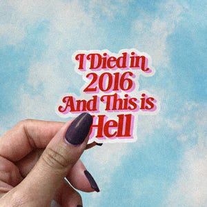 May include: A sticker with the text "I Died in 2016 And This is Hell" in red and pink lettering. The sticker is held against a blue and white cloudy background.