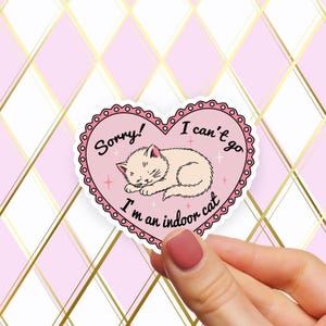 May include: A pink heart-shaped sticker featuring a sleeping cat illustration and the text "Sorry! I can't go I'm an indoor cat." The sticker has a scalloped border and is held by a hand against a pink and gold diamond pattern.