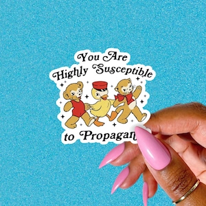 May include: A white sticker with the text "You Are Highly Susceptible to Propagan". The sticker features illustrations of a bear, a duck, and a cat walking together. The bear wears a red outfit, the duck wears sunglasses, and the cat wears a bow tie.