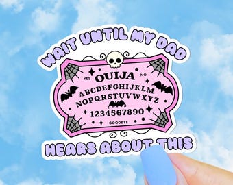 May include: A pink Ouija board sticker with the text "WAIT UNTIL MY DAD HEARS ABOUT THIS." The sticker features a skull, bats, spiderwebs, and the alphabet. The background is a blue sky with clouds.