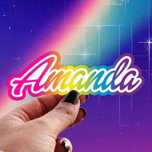 May include: A rainbow-colored sticker with the name "Amanda" written in white letters. The sticker is being held by a hand against a purple and blue background with white stars.