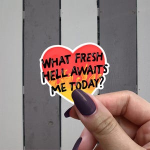 May include: A heart-shaped sticker with the text "WHAT FRESH HELL AWAITS ME TODAY?" in black letters. The heart has a red top and a yellow bottom with orange flames. The sticker is held up by a hand with purple nail polish.