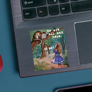 May include: A sticker featuring an illustration from Alice in Wonderland. The image shows a cat on a tree branch with the text "We are all mad here." A girl in a blue dress is standing below, with the text "At me?"