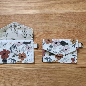 May include: Two floral print wallets. One is open, revealing a cream-colored interior with a floral pattern. The other is closed. Both wallets have a cream background with brown, pink, and green flowers.