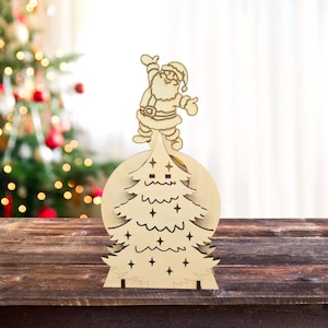 Può includere: Un ornamento per albero di Natale in legno con una figura di Babbo Natale in cima. L'ornamento è realizzato in legno marrone chiaro e presenta un design a taglio di un albero di Natale con stelle. L'ornamento è su un tavolo di legno.
