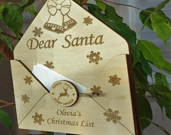 Könnte beinhalten: Holzbriefumschlag mit einem Schlitz für Briefe an den Weihnachtsmann. Der Umschlag ist mit Schneeflocken und einer Glocke verziert. Der Umschlag trägt den Text "Lieber Weihnachtsmann" und "Olivias Weihnachtsliste". Es gibt einen kleinen Holzkreis mit einem Rentier und dem Text "Nordpol Postamt" darauf.