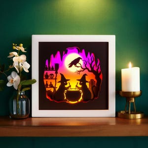 May include: A framed Halloween-themed art piece featuring silhouettes of witches, a cauldron, and a raven against a moonlit sky. The artwork is illuminated with orange and purple hues, creating a spooky ambiance. A lit candle and a vase of flowers are also present.