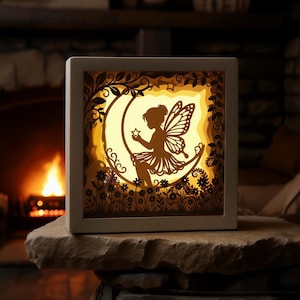 Op de afbeelding: Decoratieve, verlichte feeënlichtbox. De box toont een silhouet van een fee op een maan, die een ster vasthoudt. Het ontwerp is gelaagd met bloemen en bladeren en wordt van binnenuit verlicht, wat een warme gloed creëert.