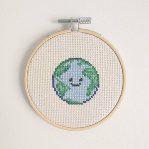 Op de afbeelding: Een handgestikte borduurring met een glimlachend cartoon-aardeontwerp. De aarde is blauw en groen op een witte achtergrond. De ring is gemaakt van natuurlijk hout.