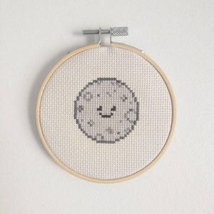 Puede incluir: Una luna bordada en gris y blanco con una cara sonriente en un aro de bordado de madera.