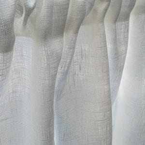 May include: A white curtain made of a semi-sheer, woven fabric with a visible textured grain. The curtain features a rod pocket header at the top, creating soft, vertical folds that allow light to pass through the material's loose weave.
