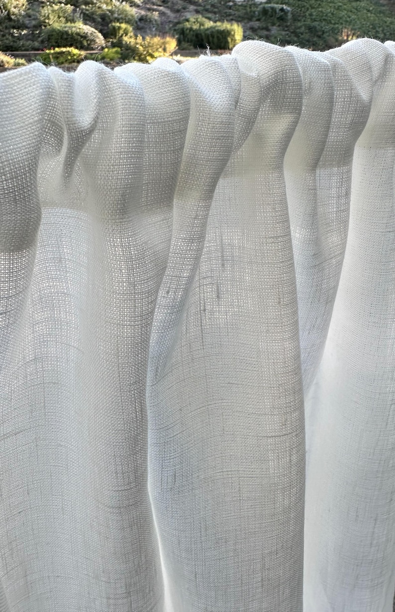 May include: A white curtain made of a semi-sheer, woven fabric with a visible textured grain. The curtain features a rod pocket header at the top, creating soft, vertical folds that allow light to pass through the material's loose weave.