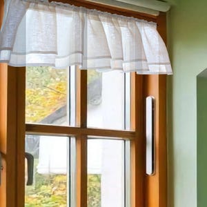 May include: A white valance curtain with a light gray border is hung above a wooden window. The window has a view of a yard with yellow and green foliage. The window frame is a dark brown color.