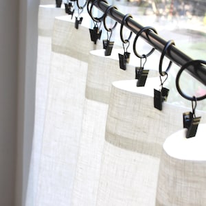 May include: A set of off-white, textured fabric curtains hanging from a black metal rod. The curtains are attached to the rod using black metal rings that feature small, integrated clips that grip the top edge of the fabric securely.