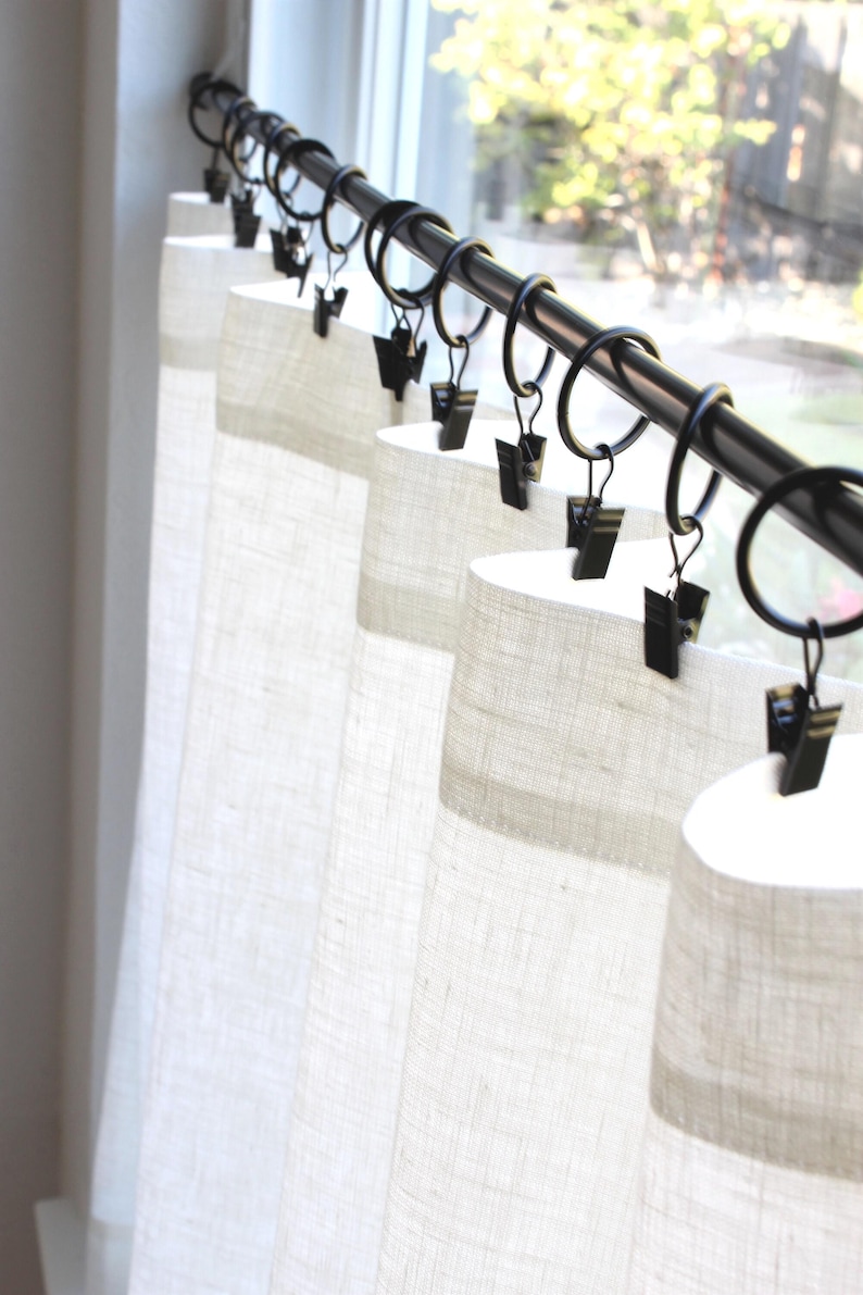 May include: A set of off-white, textured fabric curtains hanging from a black metal rod. The curtains are attached to the rod using black metal rings that feature small, integrated clips that grip the top edge of the fabric securely.