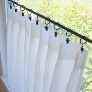 May include: White sheer curtains with pleats hanging from a black curtain rod with black clips.