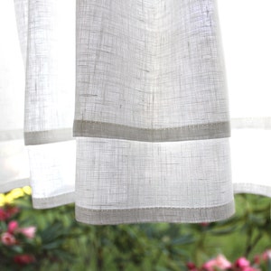 Pode incluir: Cortinas brancas e transparentes com um padr&atilde;o texturizado sutil e uma fina bainha branca ao longo da borda inferior. As cortinas est&atilde;o penduradas em uma janela e a luz brilha atrav&eacute;s delas.