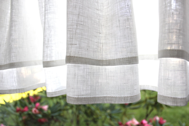 Pode incluir: Cortinas brancas e transparentes com um padr&atilde;o texturizado sutil e uma fina bainha branca ao longo da borda inferior. As cortinas est&atilde;o penduradas em uma janela e a luz brilha atrav&eacute;s delas.