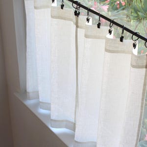 May include: A set of off-white, semi-sheer curtains hanging from a black metal rod. The curtains are attached to the rod using black metal rings with integrated clips, creating vertical folds along the fabric panels that cover the window.