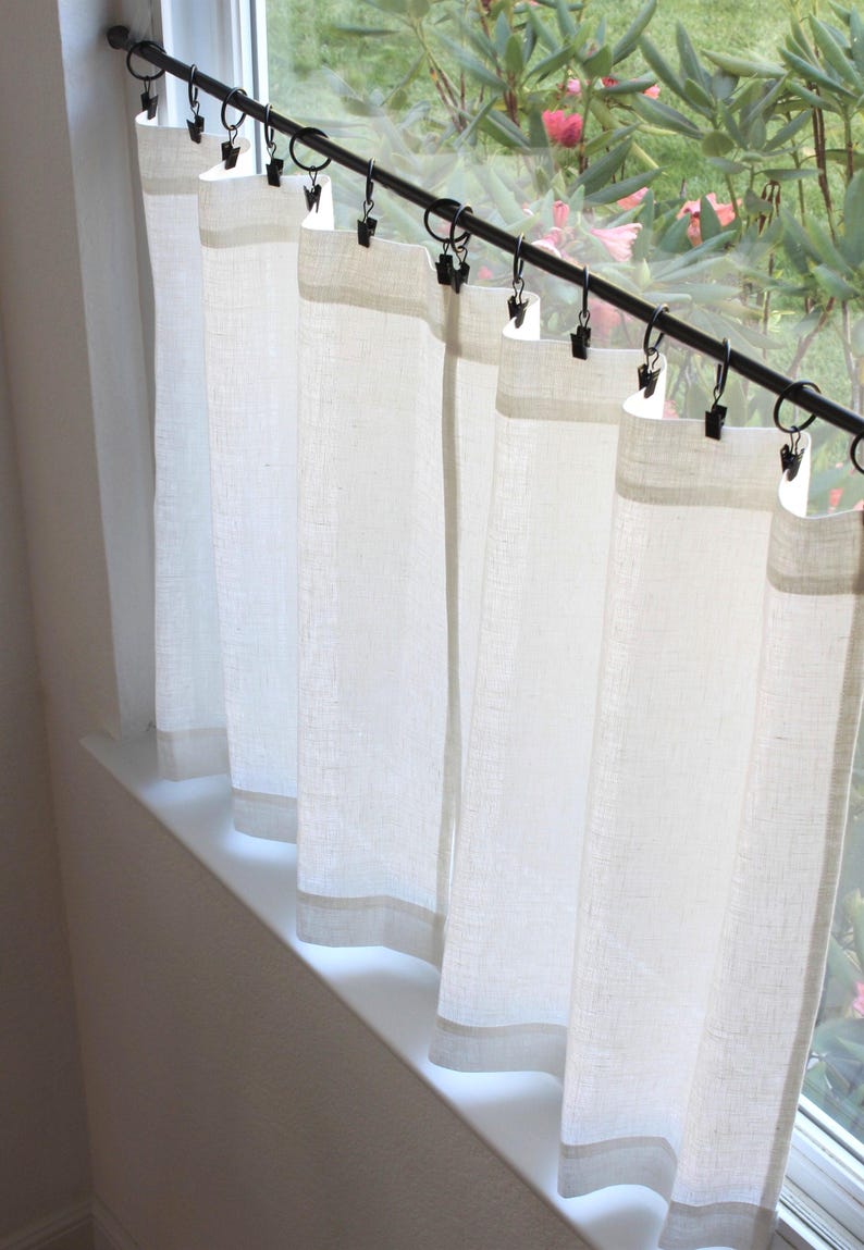 May include: A set of off-white, semi-sheer curtains hanging from a black metal rod. The curtains are attached to the rod using black metal rings with integrated clips, creating vertical folds along the fabric panels that cover the window.