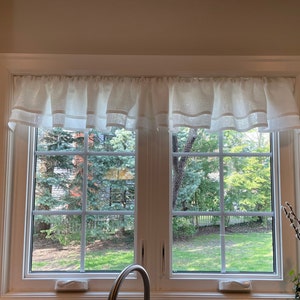 Pode incluir: Uma valance de linho branco com uma borda franjada pendurada sobre uma janela dupla com vista para um gramado verde e &aacute;rvores.