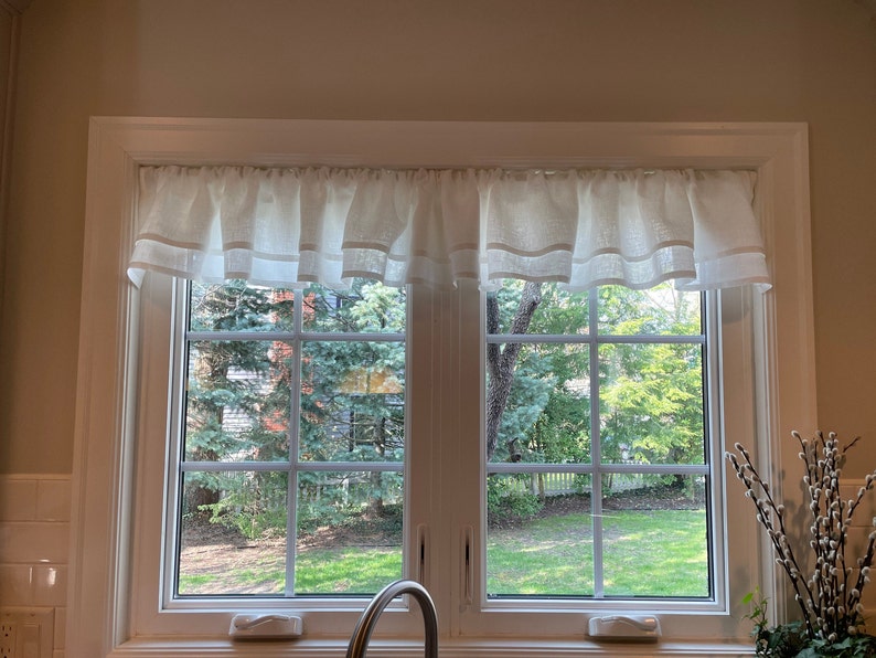 Pode incluir: Uma valance de linho branco com uma borda franjada pendurada sobre uma janela dupla com vista para um gramado verde e &aacute;rvores.