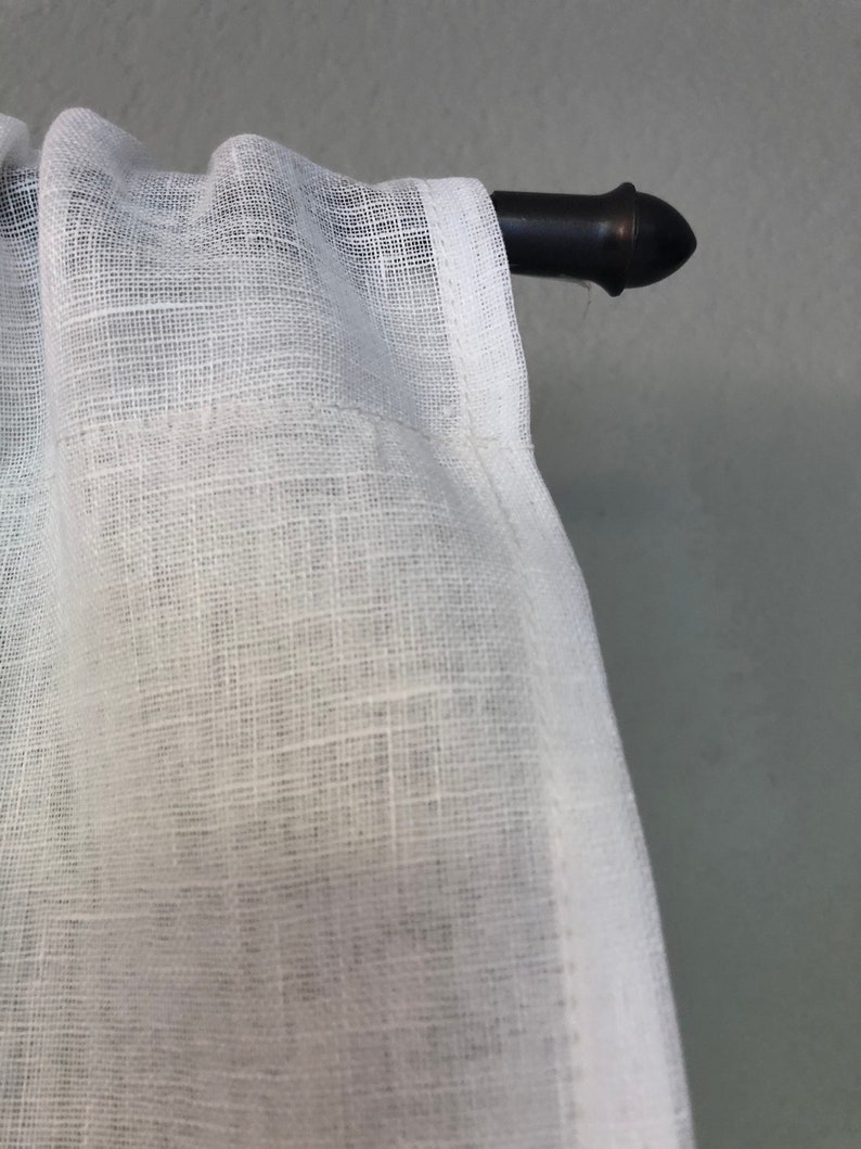 Pode incluir: Cortina branca transparente com textura de linho pendurada em uma haste de cortina de metal preto com uma ponta decorativa.