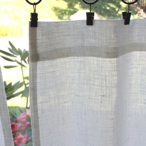May include: A semi-sheer white curtain panel with a visible woven texture, suspended from a black metal rod by three black circular rings with attached clips. The fabric has a light, airy appearance, allowing natural light to pass through.