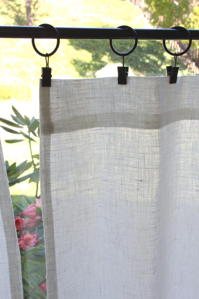 May include: A semi-sheer white curtain panel with a visible woven texture, suspended from a black metal rod by three black circular rings with attached clips. The fabric has a light, airy appearance, allowing natural light to pass through.