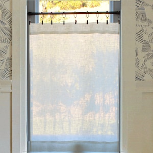 May include: A semi-sheer, off-white linen curtain panel hangs from a dark metal rod using a series of black clip rings. The rectangular fabric features a visible weave texture and covers the centre portion of a window, allowing light to pass through.