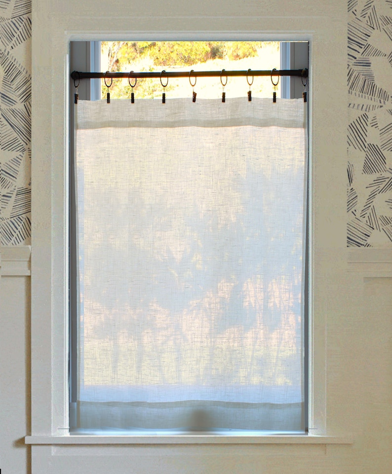 May include: A semi-sheer, off-white linen curtain panel hangs from a dark metal rod using a series of black clip rings. The rectangular fabric features a visible weave texture and covers the centre portion of a window, allowing light to pass through.